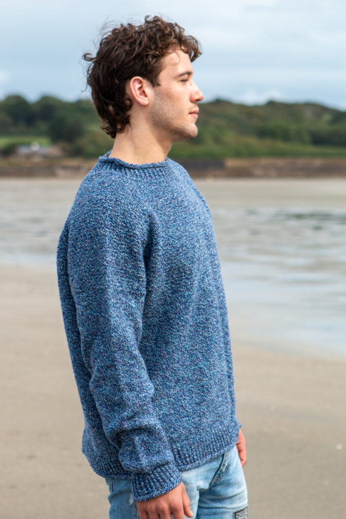 Man wearing a handknit sweater designed by carol feller in a blue marled Irish yarn. 